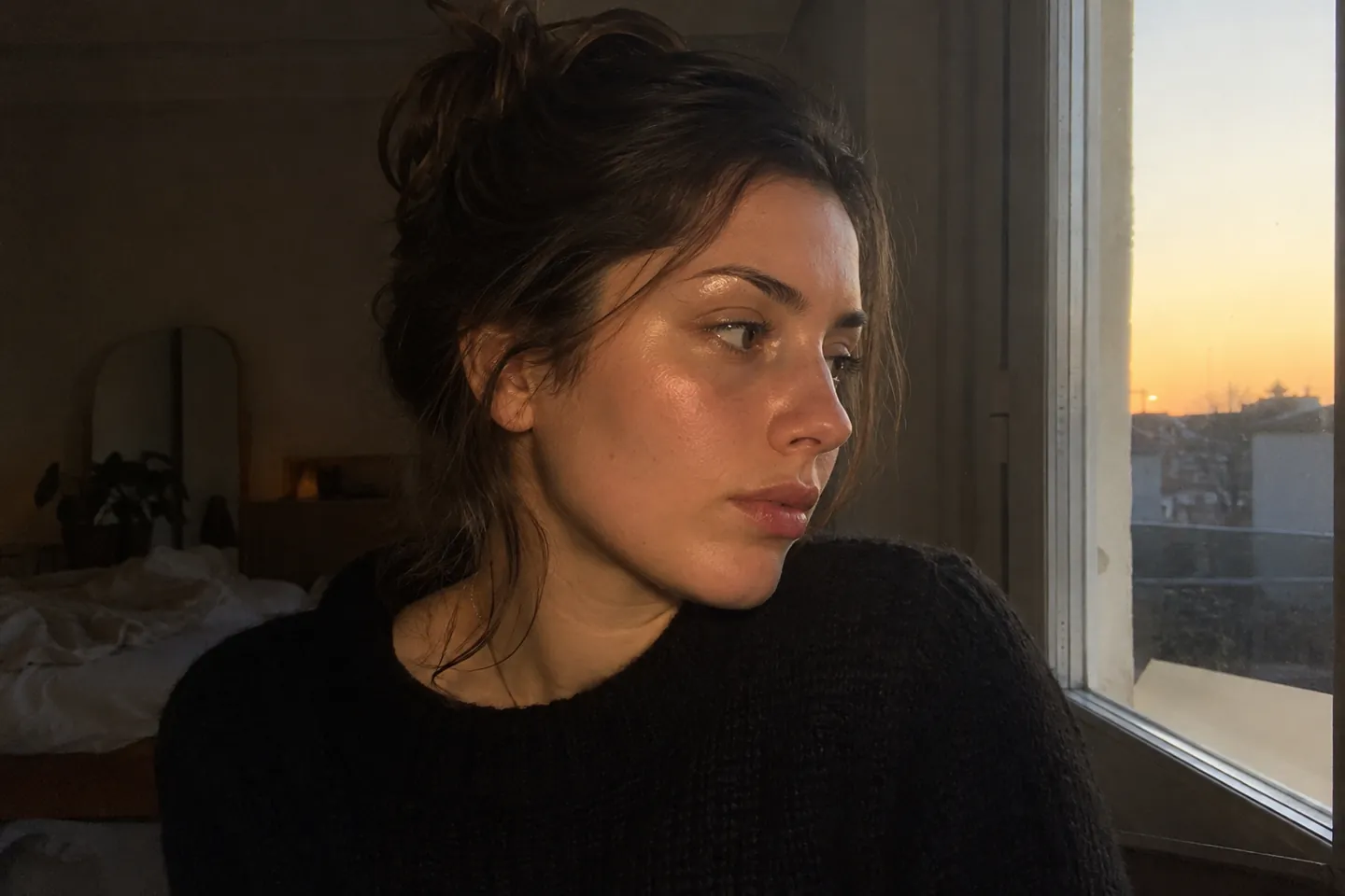 A woman in a dim room with a gold shimmer on her cheekbones, looking away